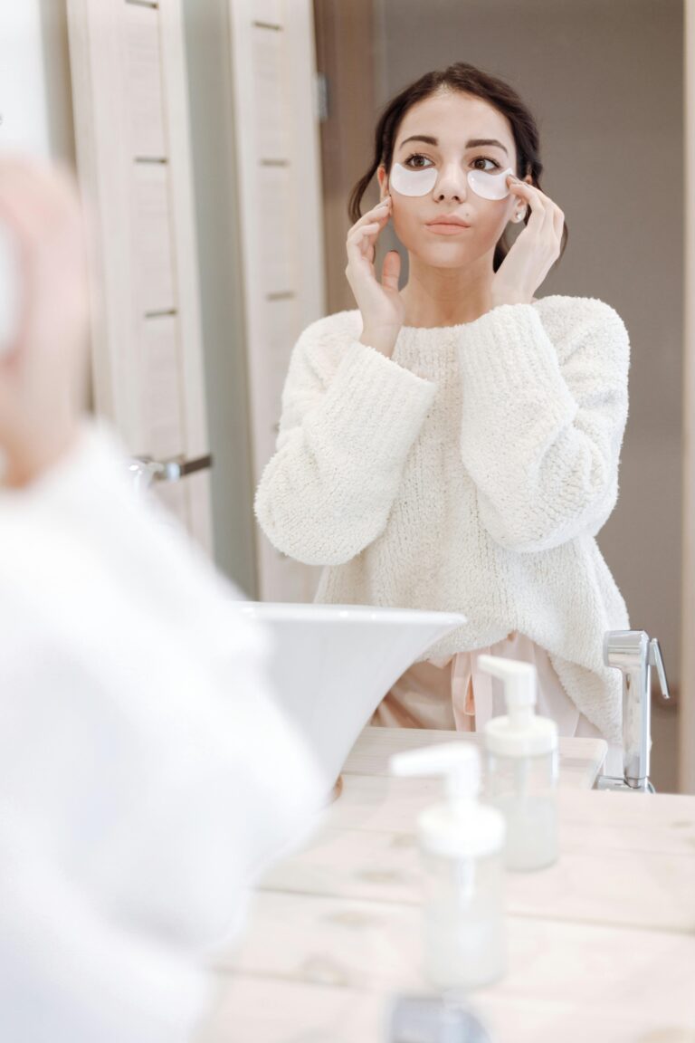 Woman in white sweater applying under eye masks while looking in mirror, focusing on skincare routine.
